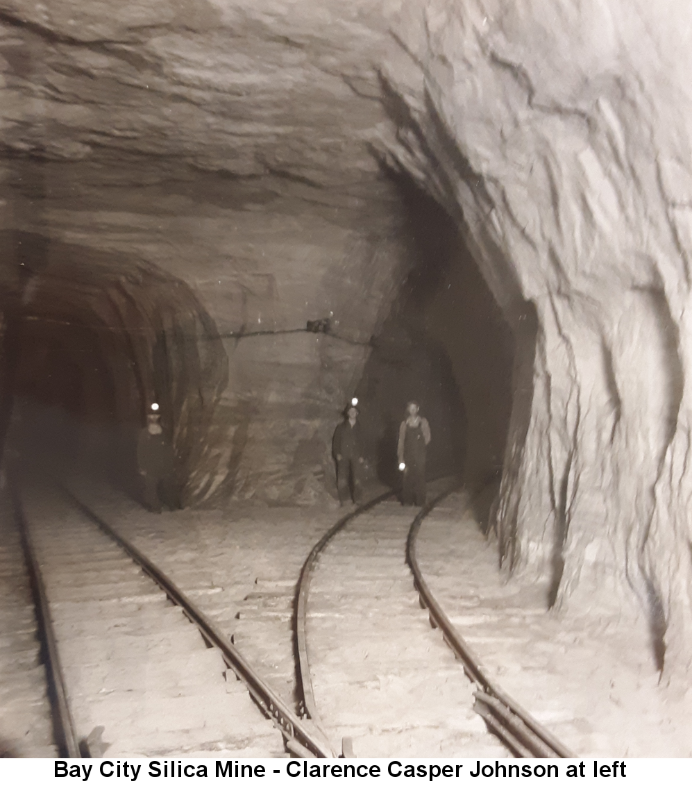 Black and white photo of two mine tunnels branching off with railroad tracks running into them; Clarence Casper Johnson, in overalls, his helmet light a bright round dot, stands at the entrance to the left tunnel; two other miners stand in the right tunnel.